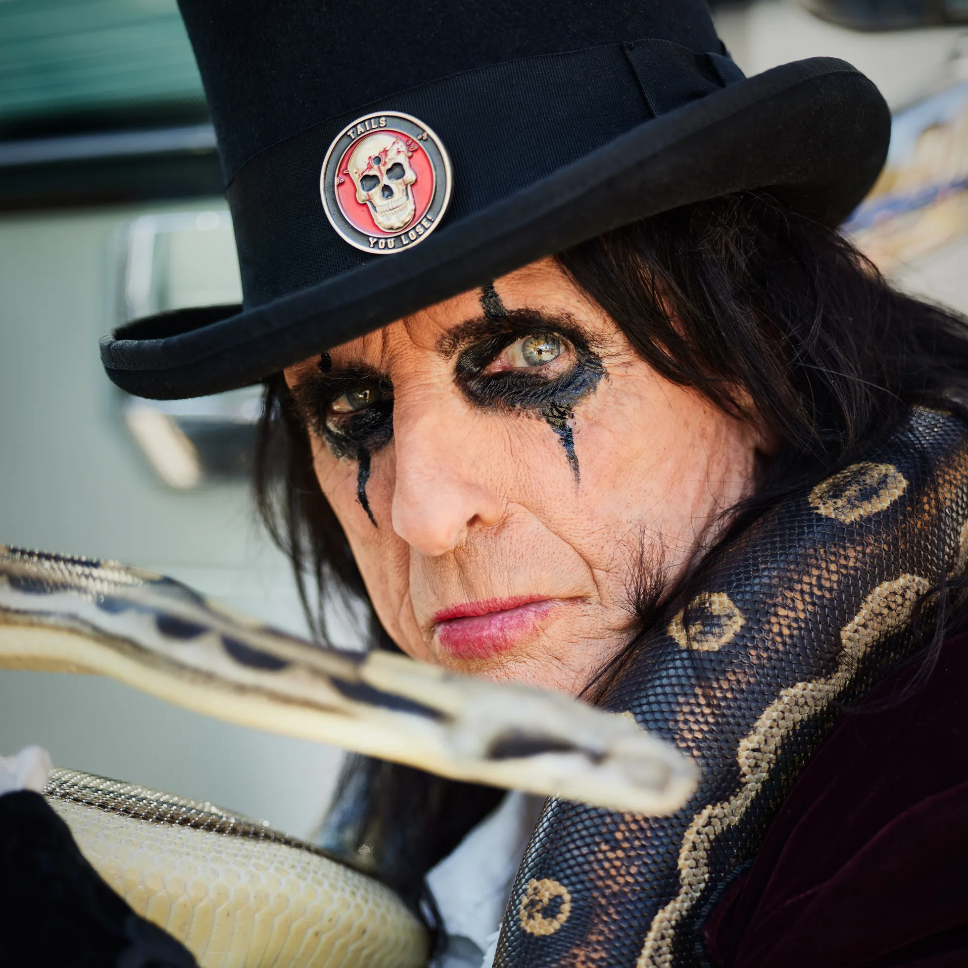 Alice Cooper wearing a black top hat with a 'TAILS YOU LOSE' skull pin, heavy black eye makeup, holding a large python snake around his neck.