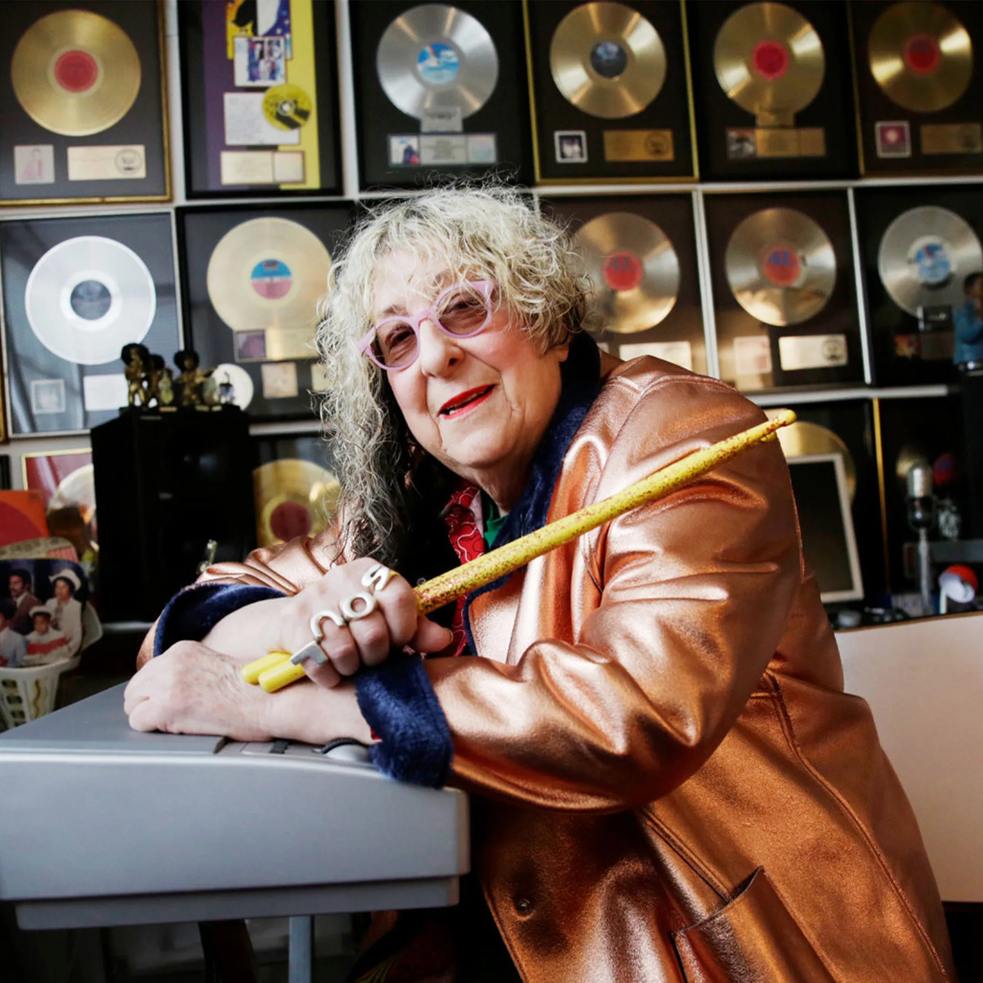 Allee Willis, legendary songwriter, smiling in a shiny copper jacket and pink glasses, holding a glittery drumstick with 'SOUL' rings on her fingers, in front of a wall of gold and platinum records.