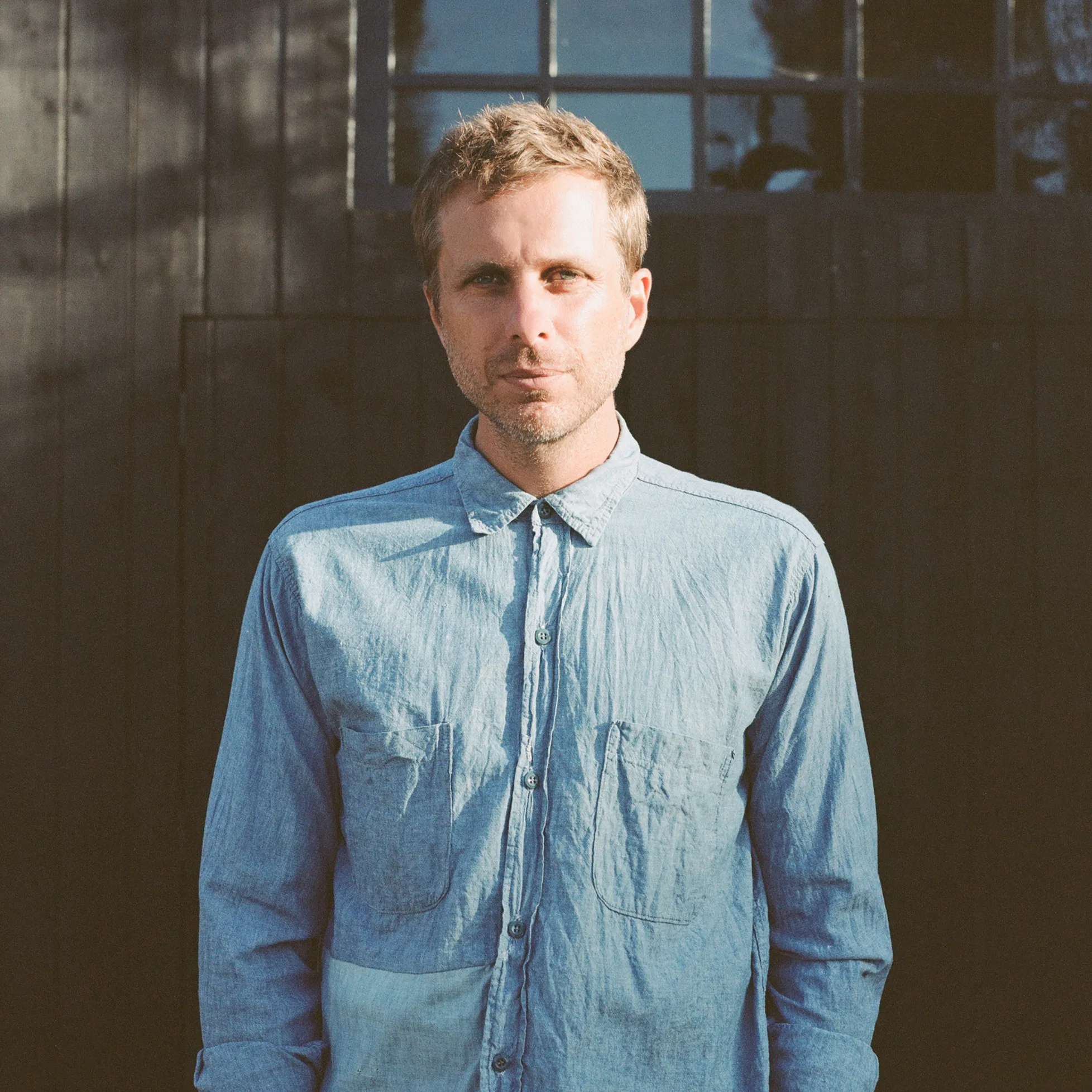 Aaron Bruno of AWOLNATION – official portrait of the lead singer in a blue denim shirt standing in front of a dark wooden building.