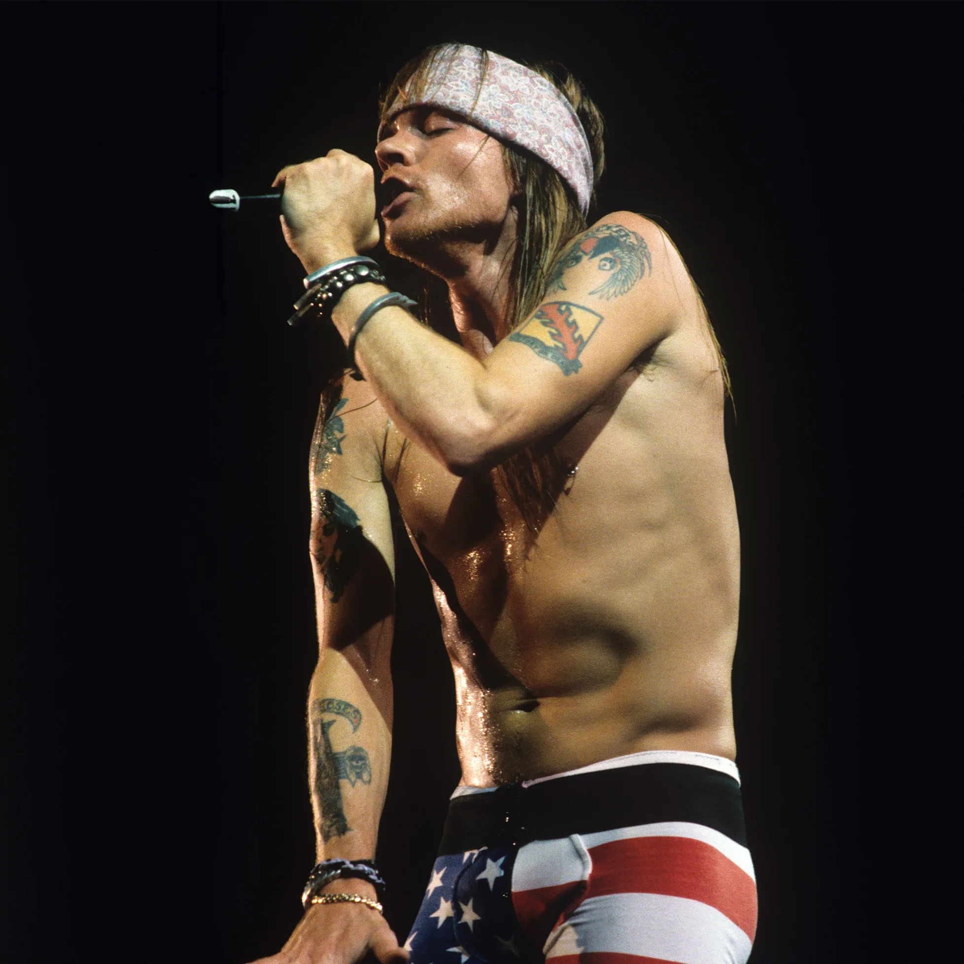 Axl Rose of Guns N' Roses performing live on stage, shirtless with bandana, tattoos visible, singing into a microphone while wearing American flag pants.
