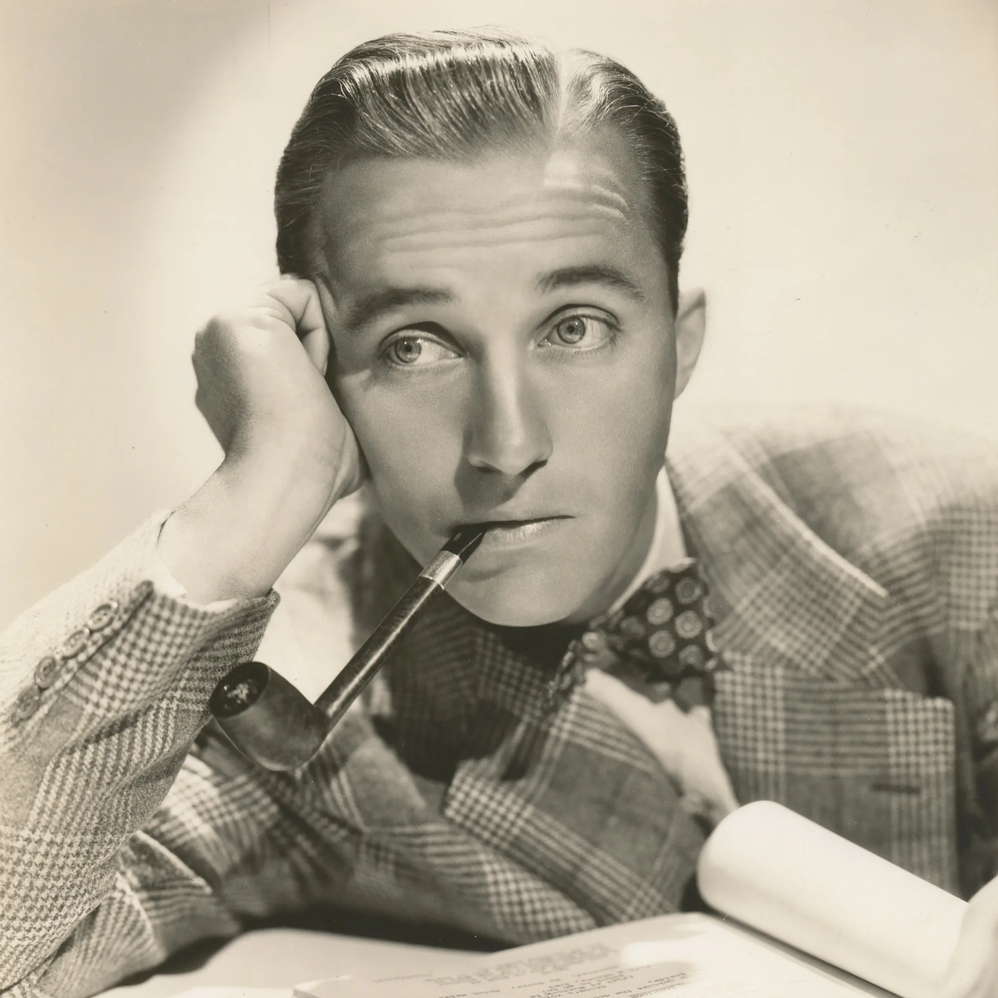 Bing Crosby classic black and white portrait, holding a pipe in his mouth with thoughtful expression, wearing a plaid jacket and bow tie.