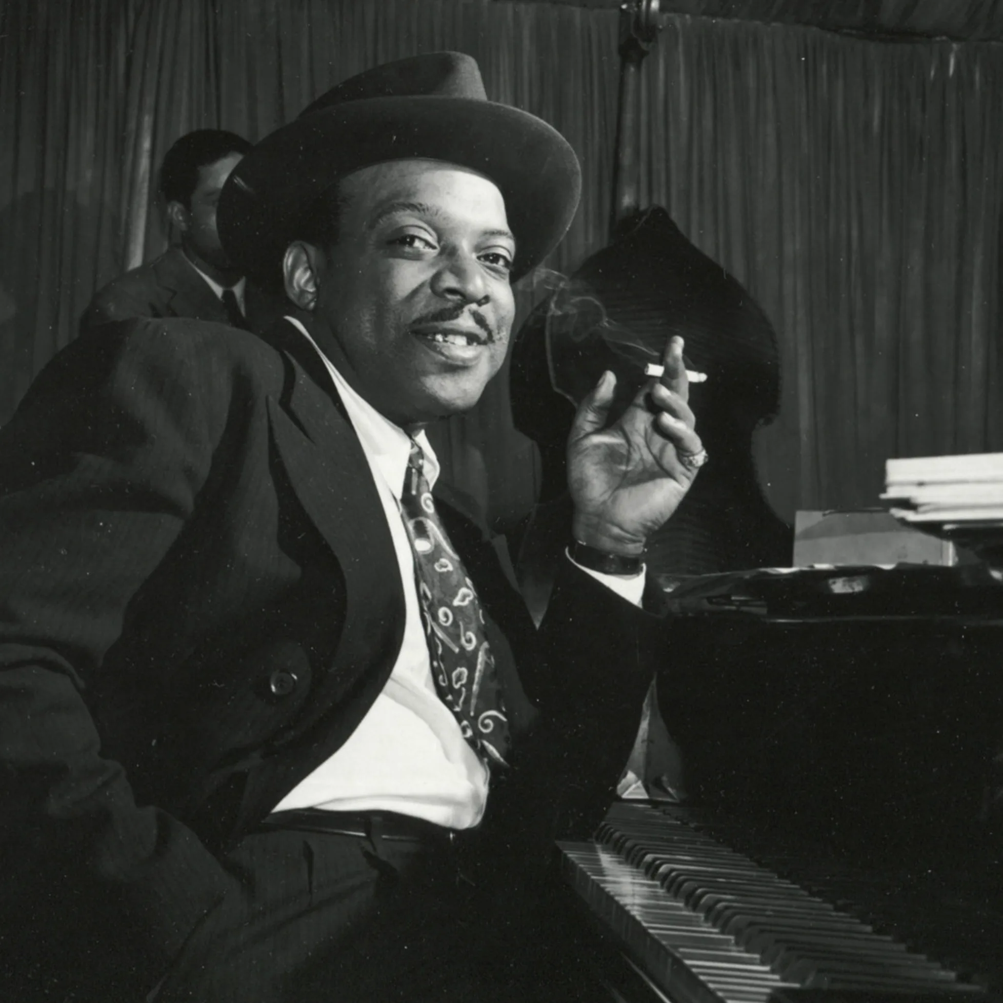 Count Basie in fedora hat playing piano with cigarette.