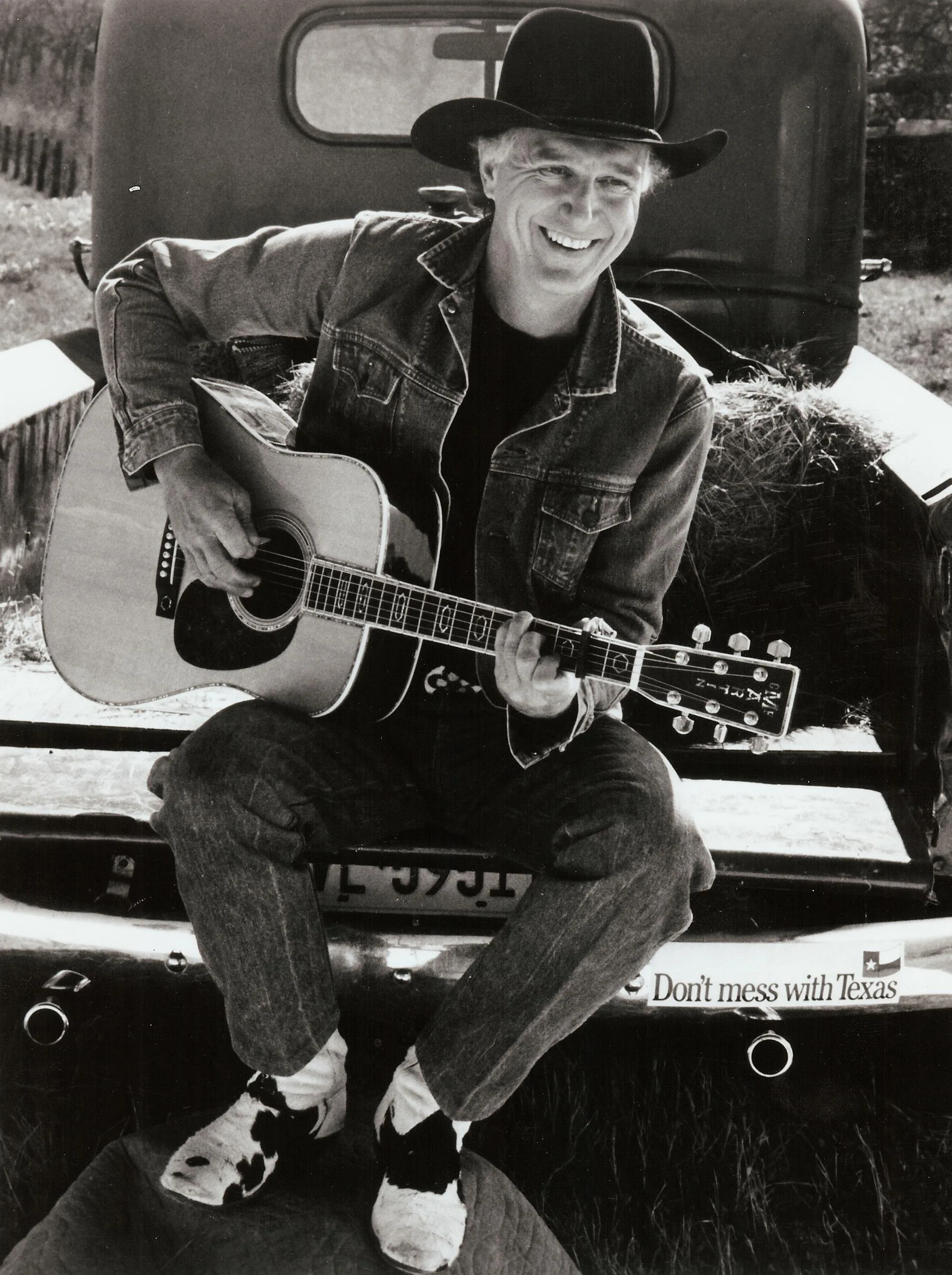 A black and white photo of Jerry Jeff Walker sitting on the back of a pickup truck, playing guitar.