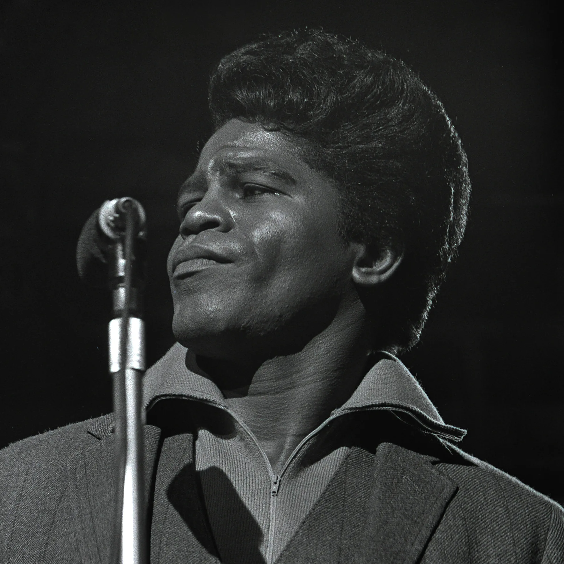 James Brown performing live on stage, iconic close-up portrait singing passionately into a vintage microphone, wearing a zip-up shirt and jacket.