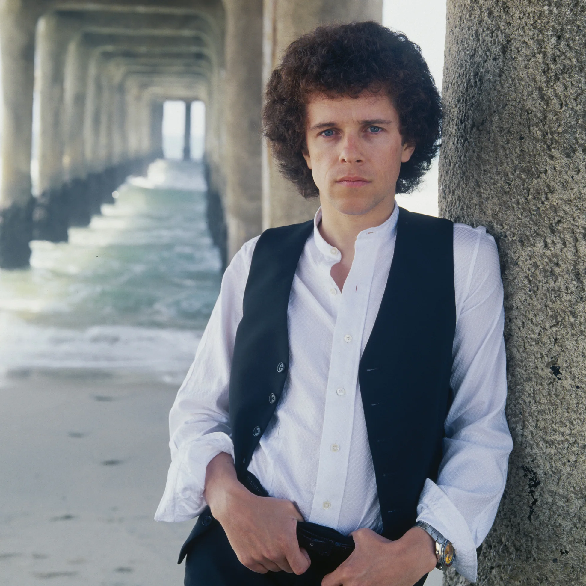 Leo Sayer leaning against a pier with curly hair, wearing a black vest and white shirt.