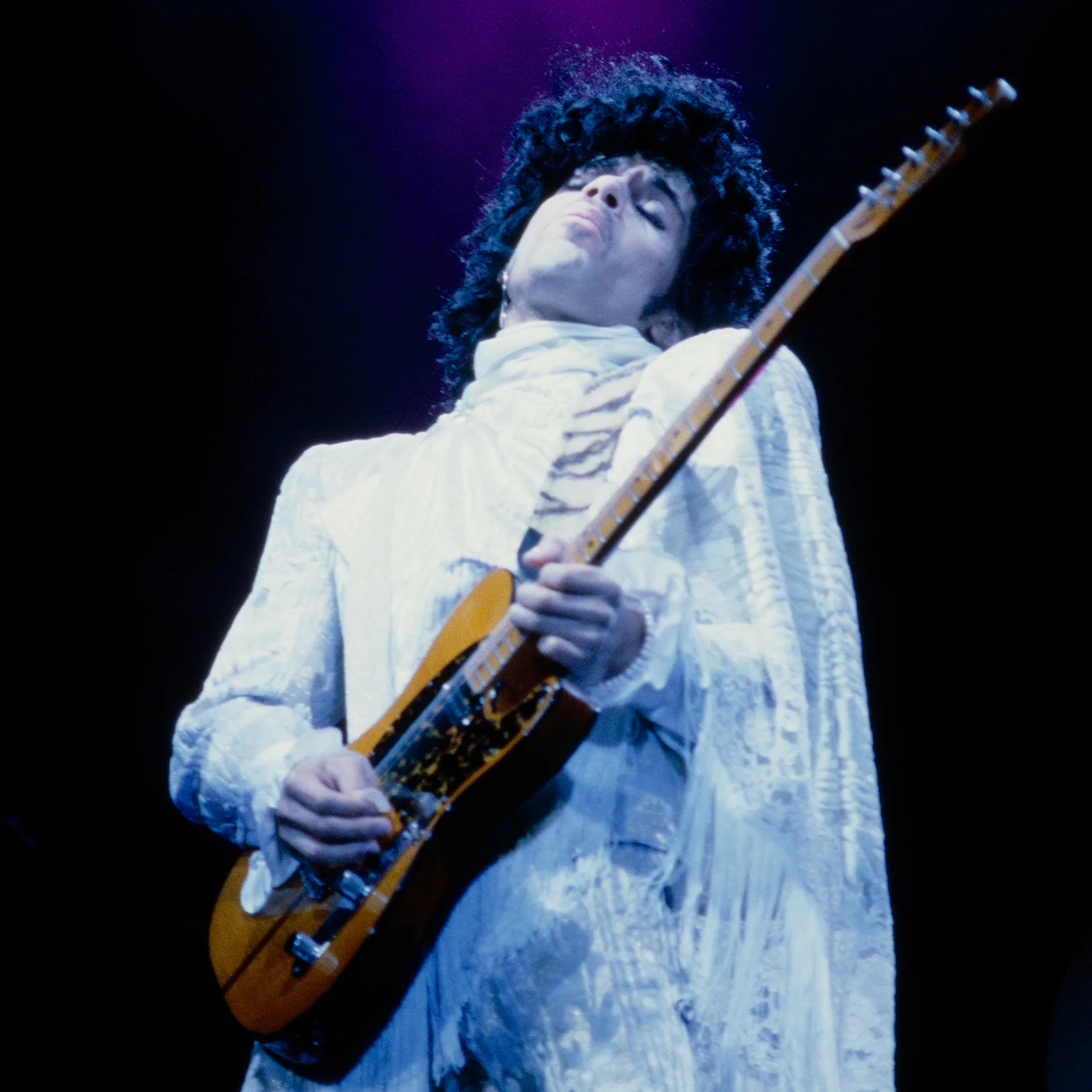 Prince performing live in white outfit, playing electric guitar under purple stage lights