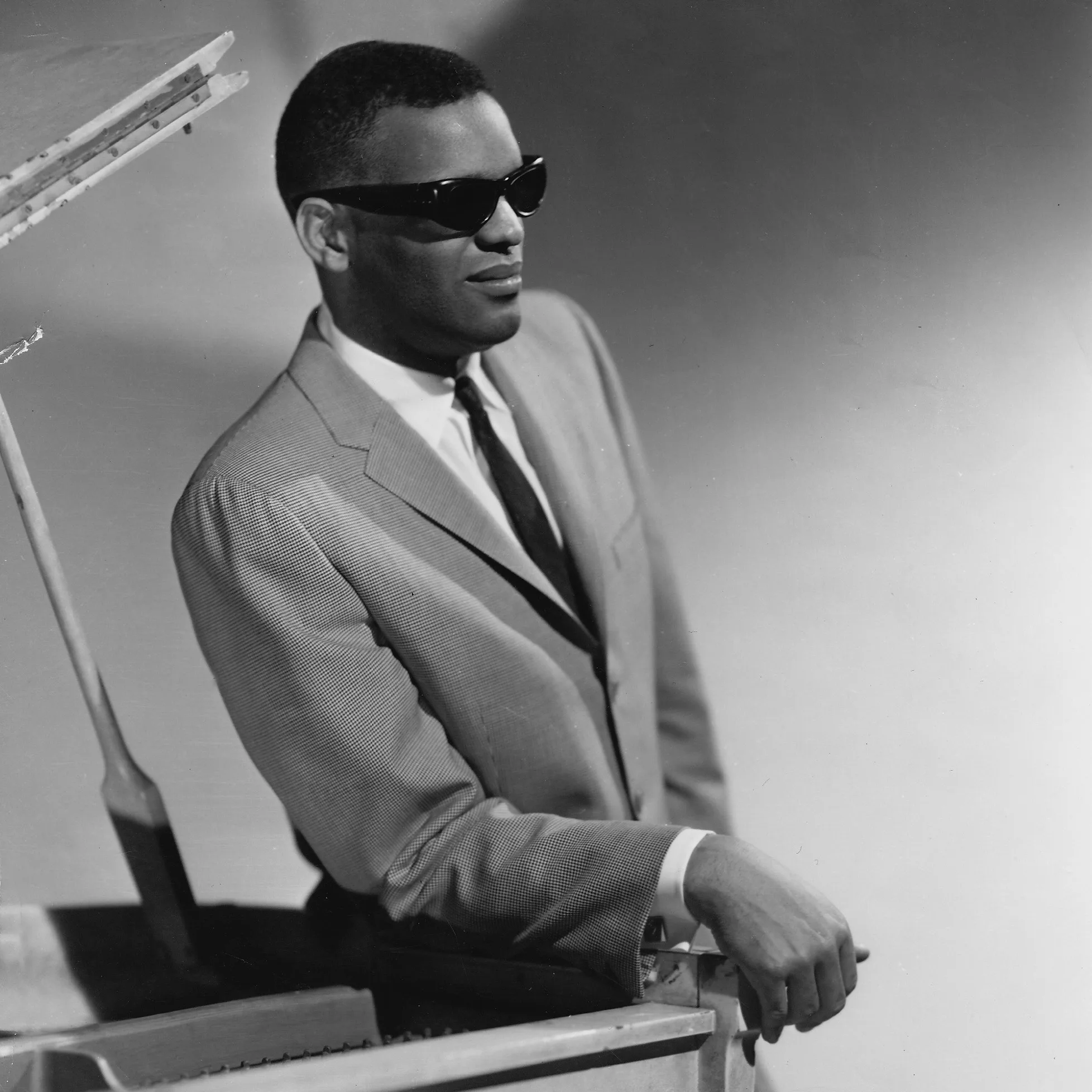 Ray Charles classic black and white portrait at the piano, wearing signature sunglasses and suit, leaning on grand piano.