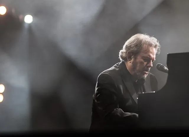 Jimmy Webb performing at a piano with stage lights and smoke around him.