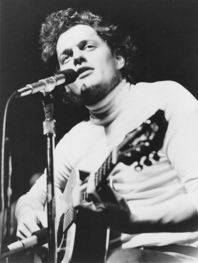 Harry Chapin singing and playing guitar on stage in a black and white photo.