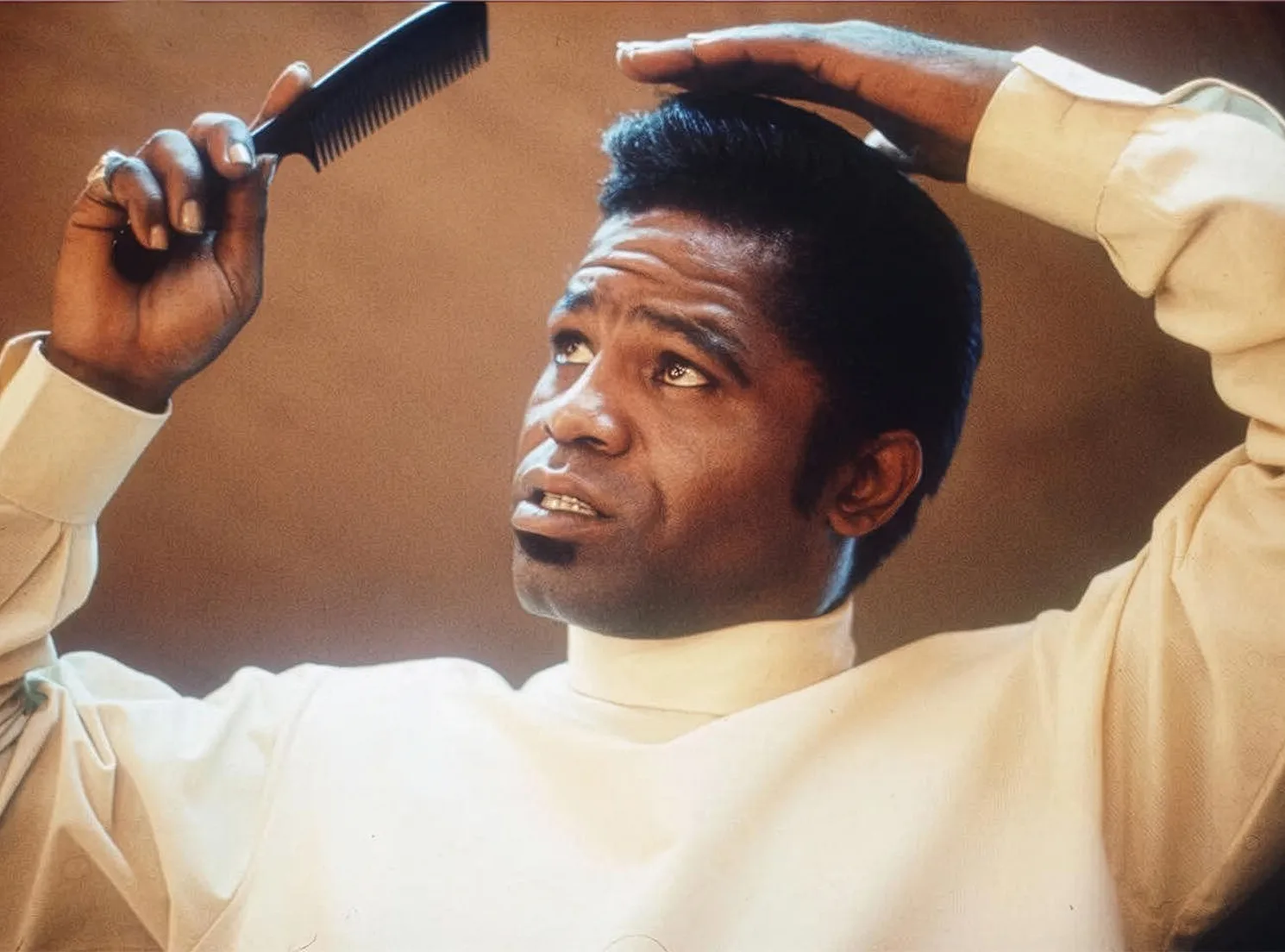 James Brown styling his iconic pompadour hairstyle with a comb, wearing a white turtleneck.